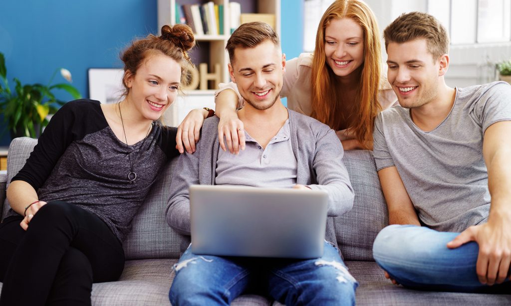 Neljä hymyilevää nuorta aikuista katsoo tietokoneen ruutua sohvan ympärillä. 
