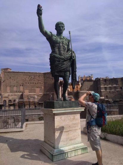 Kädellään ylöspäin viittaavan keisarin patsas Roomassa. Taustalla näkyy kaivauksissa esiinotettuja kivimuureja.